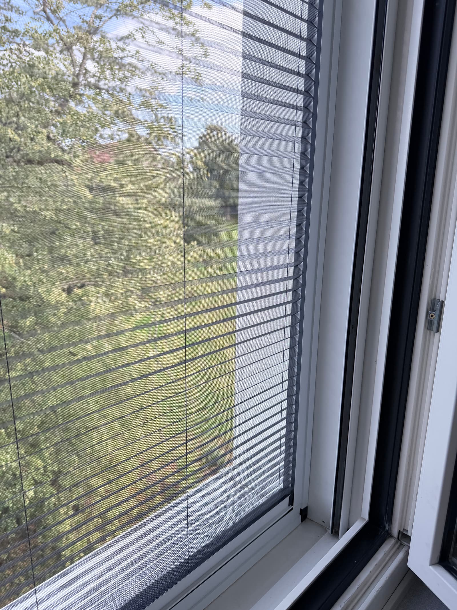 Nahaufnahme des Plissees: Links liegt das Gewebe gespannt vor der Scheibe, rechts sind die Falten zusammengeschoben. Dahinter Baum und Wiese.