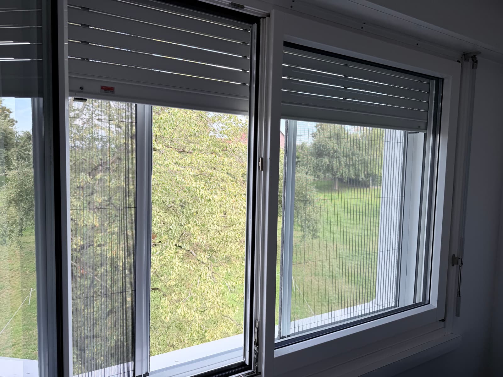 Zwei nebeneinanderliegende Fenster von innen, an beiden ist ein Insektenschutz-Plissee montiert; am linken ist es teilweise zugezogen. Über beiden sind die Lamellenstoren hochgezogen.